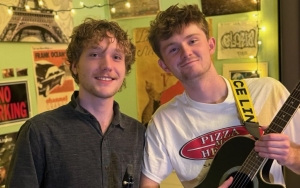 Two young men stand indoors. One holds a guitar, and they are surrounded by posters and string lights.