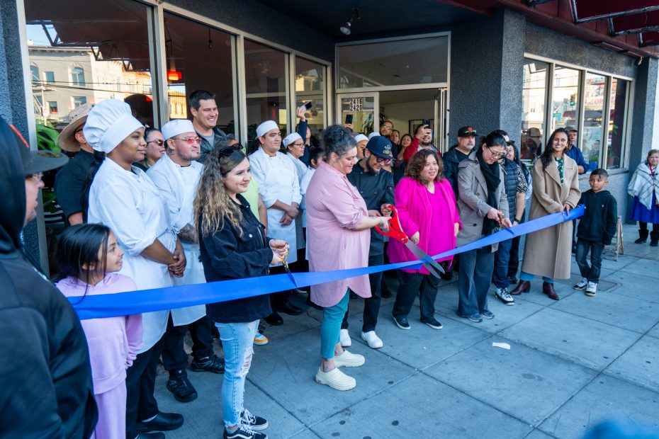 A group of people, including chefs in white uniforms, gathers outside a building for a ribbon-cutting ceremony. A woman in pink cuts the blue ribbon while others look on.