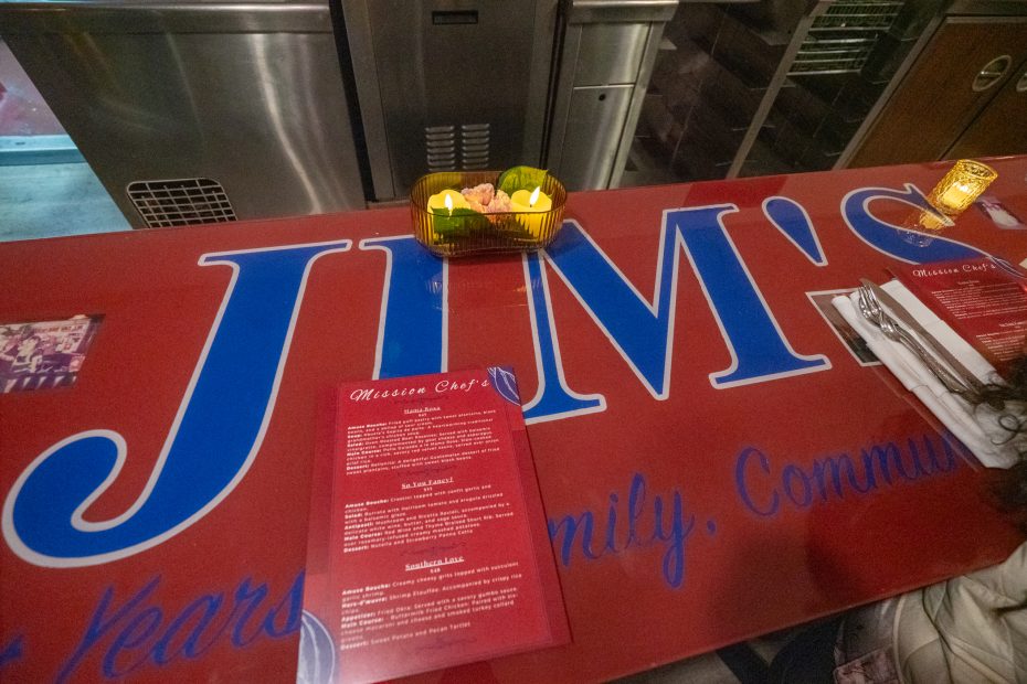 Red countertop with large blue letters spelling "JIM'S." A small green dish with lemon wedges, a candle, and red menus are displayed on the counter.