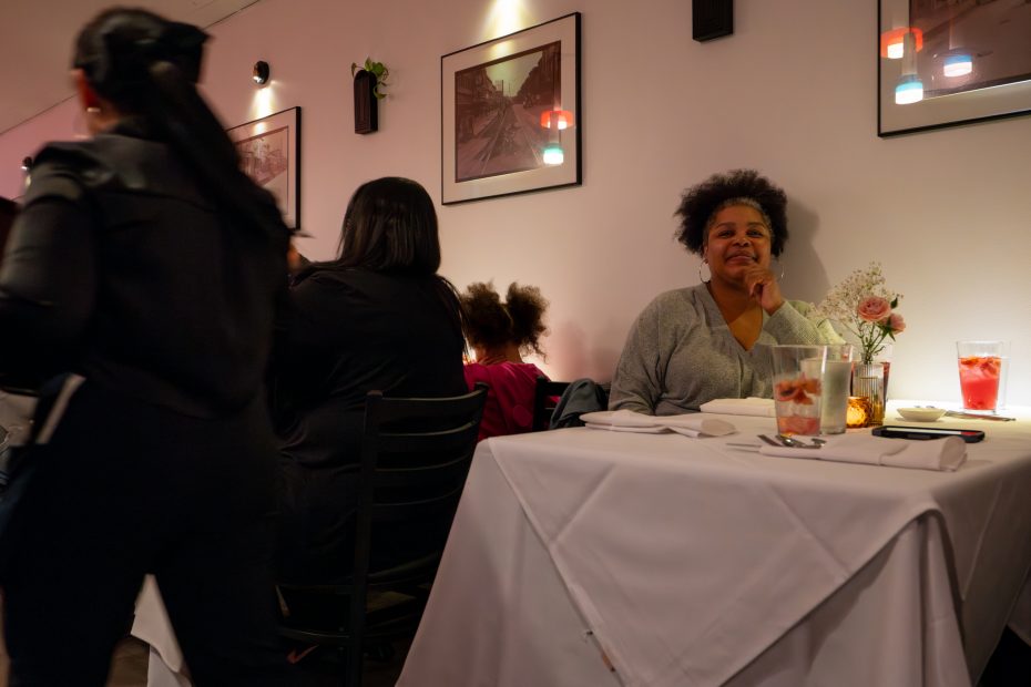 Woman sitting at a restaurant table with a drink, observing surroundings. Other diners and pictures on the wall are visible in the background.