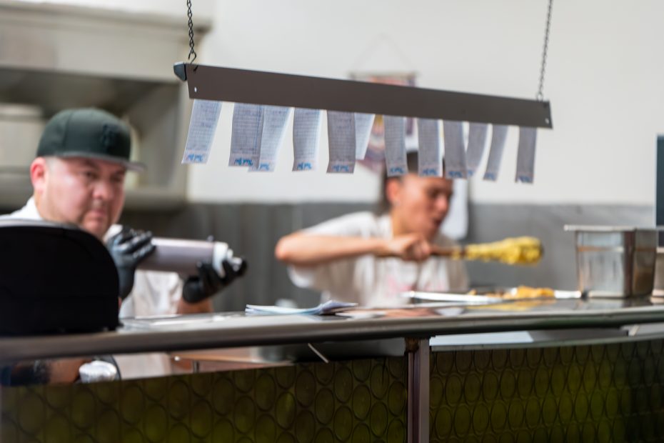 Two chefs in a kitchen work behind a counter. Cooking order tickets hang above them on a rack. One chef is preparing food, while the other is using a stainless steel container.
