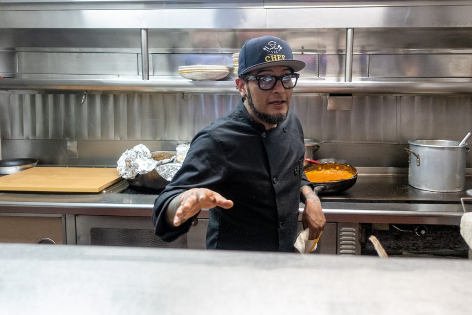 Chef in a black uniform and cap standing in a commercial kitchen with pots and foil-wrapped items in the background.