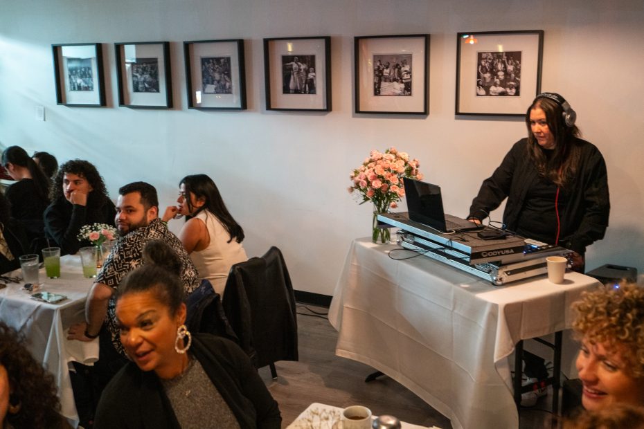 A DJ stands at a booth near a wall with framed photos, playing music for people seated at tables, some with drinks. Flowers in a vase decorate the DJ's table.