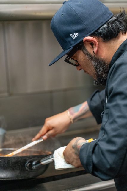 Chef in a black hat and coat cooks food in a pan on a stove, using a spatula with a towel in hand for grip.