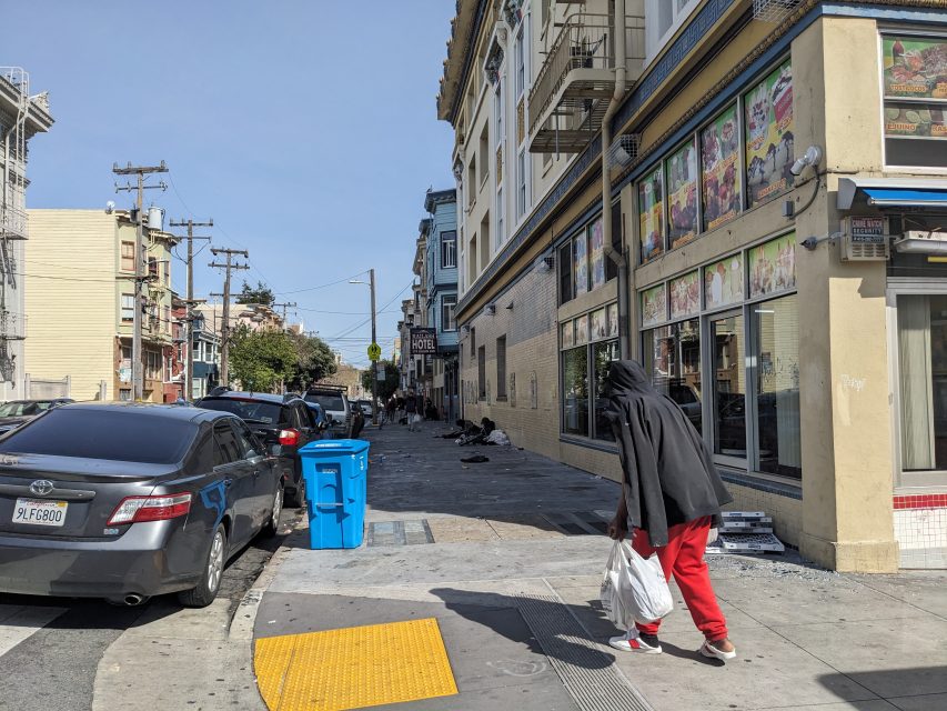 A person in a hooded jacket and red pants walks down a city street holding two plastic bags. Several parked cars and a building with colorful signs are visible.