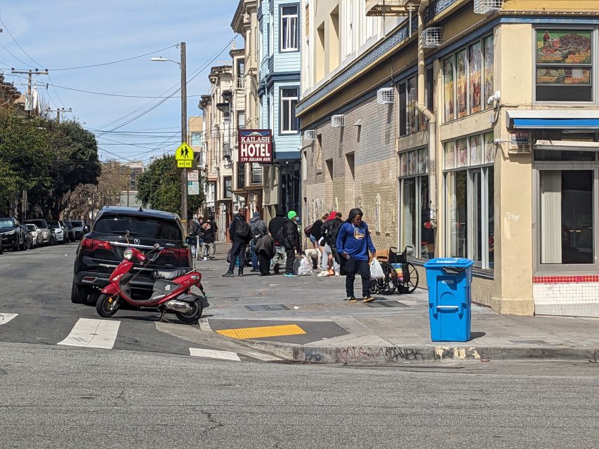 People gather on a sidewalk near a building with a "Hotel" sign. A parked car and scooter are nearby. An open blue trash bin is visible on the street corner.