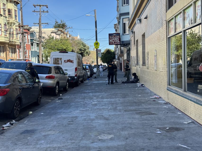 City street with parked cars and a white van labeled "Plumbing." Three people are on the sidewalk near a building with a "Hotel" sign. Debris is scattered on the ground.