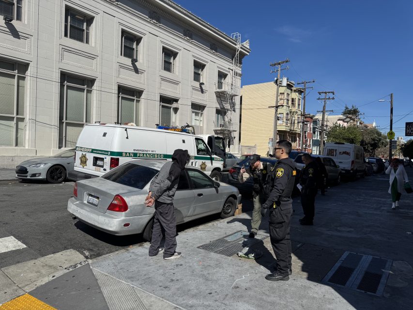 Police officers speak to individuals on a city sidewalk near parked vehicles, including a police van, beside a large building.