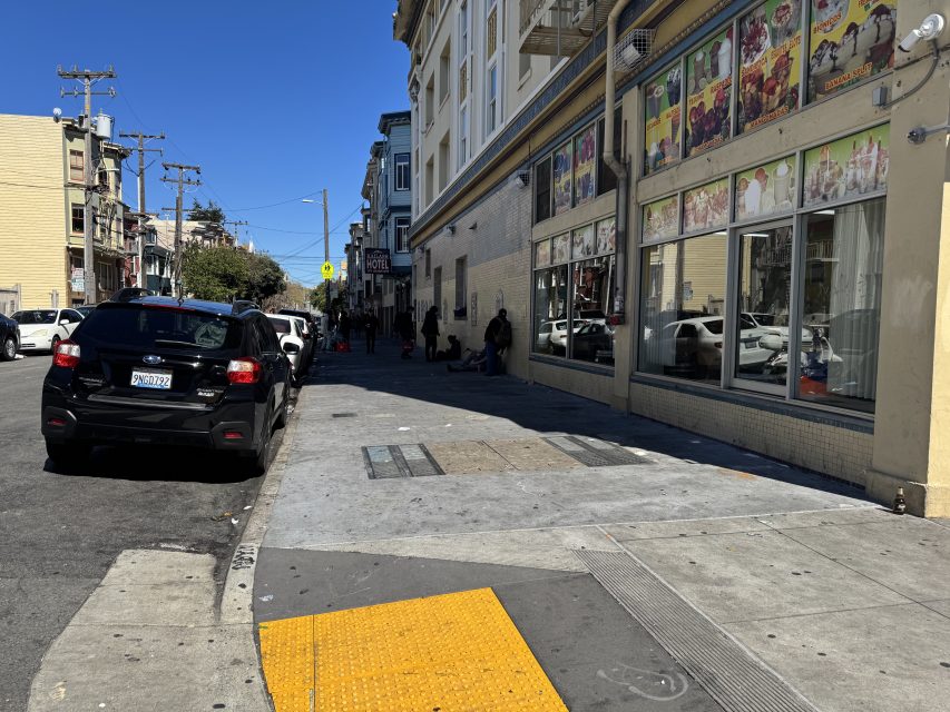 Urban street scene with parked cars, a storefront displaying colorful posters, and people gathered on the sidewalk under a clear blue sky.