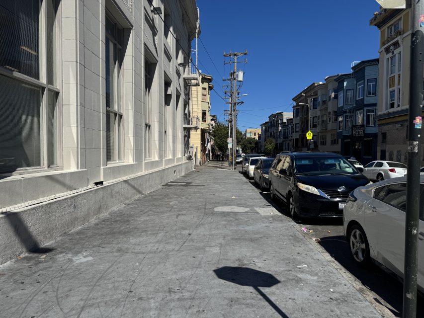 A city sidewalk with a row of parked cars on the right, buildings lining both sides, and a clear blue sky above.