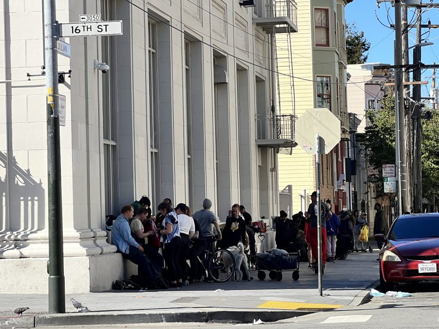 A group of people gathered outside a building on the lively 16th Street BART plaza. Some are seated, others stand amid carts and bags, while a red car is parked nearby.