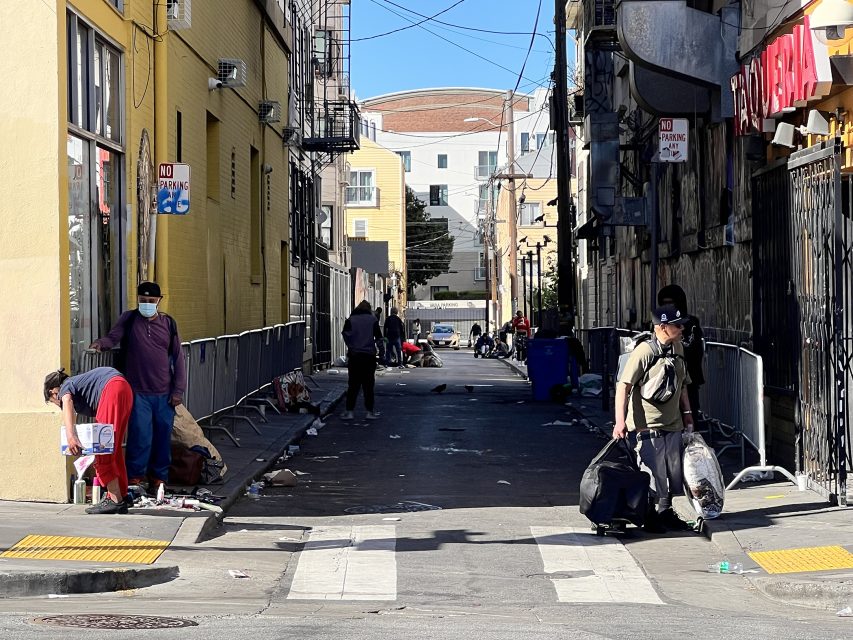 An urban alleyway near the bustling 16th Street BART plaza shows several people navigating through scattered debris. Buildings line the sides, and a "no parking" sign stands prominently, adding to the textured city scene.