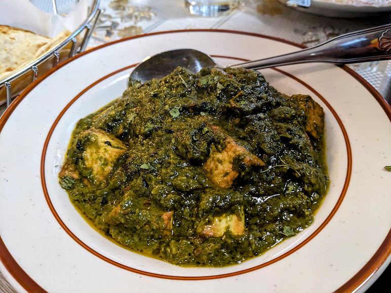 A plate of saag paneer with a serving spoon. The dish features paneer cubes in a thick, green spinach gravy on a patterned tablecloth.