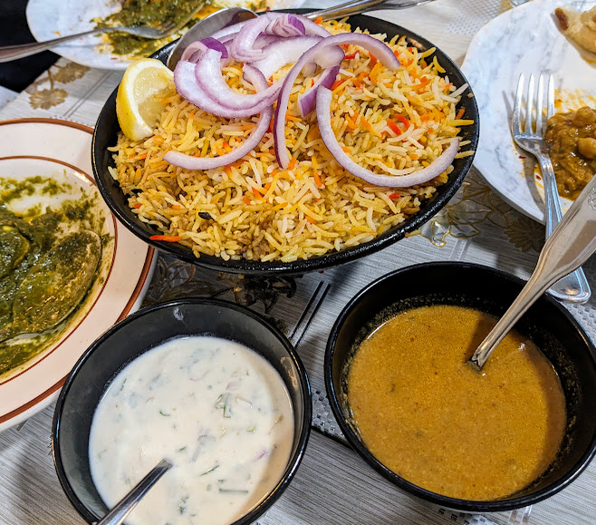 A bowl of biryani topped with onion slices and a lemon wedge, accompanied by bowls of raita and curry on a table. Other dishes are partially visible.