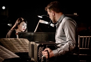 A man plays a bandoneon while seated next to a woman playing a piano. Music sheets are visible in front of them.