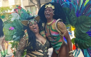 Two people in vibrant carnival costumes, one with leopard print and another with peacock-inspired feathers and sequins, smile and pose outdoors.