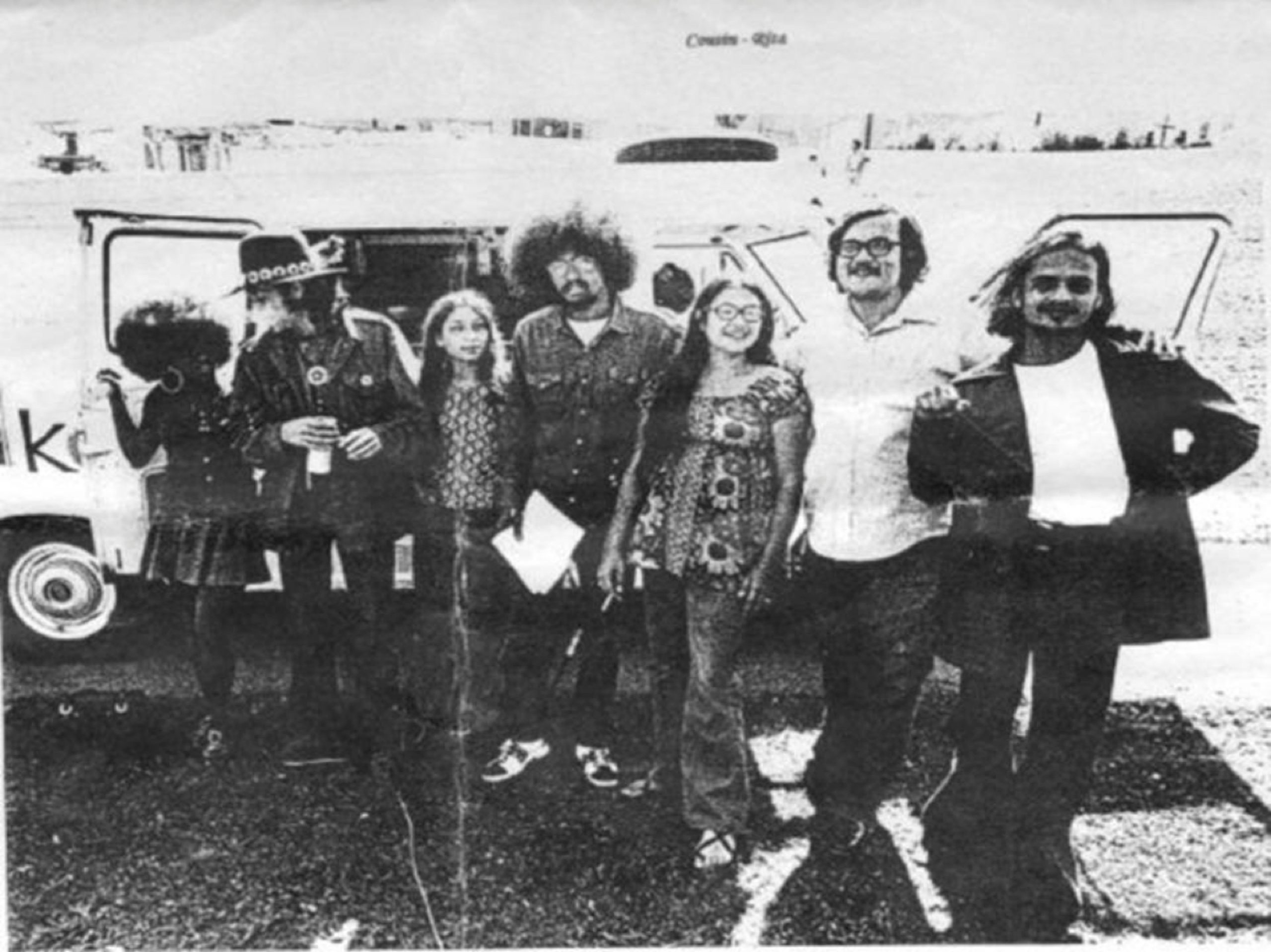 A group of seven people stand in front of an open van on a sunny day, posing together. They are casually dressed, and the background features a gravel area and blurred buildings.