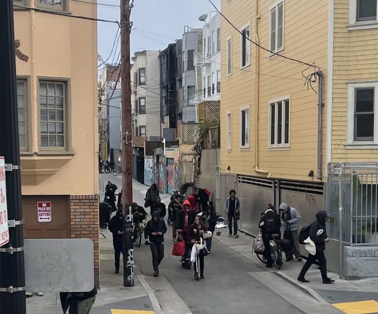 A group of people gathers in a narrow urban alley between residential buildings. Some are standing with bags, and others are moving around. Graffiti is visible on the walls.