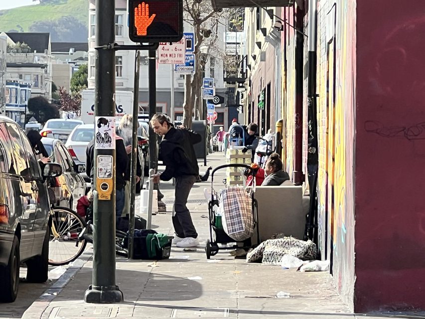 Street scene with people near a sidewalk filled with various items. Cars are parked on the left, and a pedestrian wait signal is visible. A person with a stroller is seated by a wall.