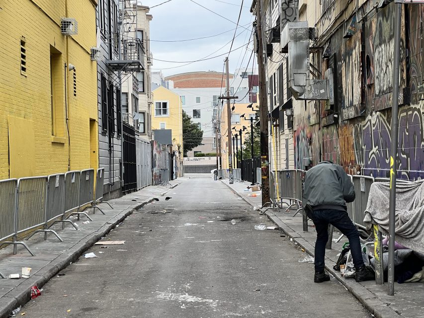 A person leans over near a wall in a narrow urban alley lined with barricades and graffiti. The alley shows scattered debris and leads to a distant street.