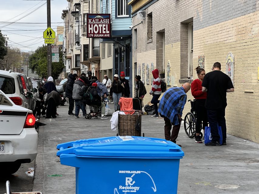 People congregate on a city sidewalk beside parked cars and a blue recycling bin. A hotel sign is visible. Some individuals are sitting or using wheelchairs.