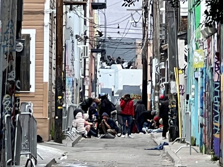 A narrow urban alley features various people gathered, surrounded by graffiti-covered walls and overhead power lines.