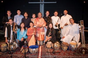 A group of musicians pose on a stage with various Indian classical instruments, including sitars and tablas, wearing a mix of traditional and modern clothing, smiling for the camera.