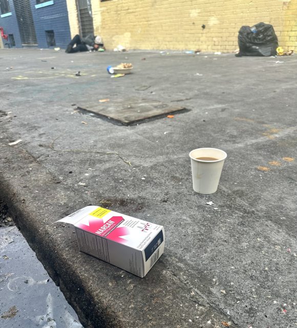 Discarded coffee cup and medication box on a littered urban sidewalk, with a person in the background sitting against a yellow brick wall.