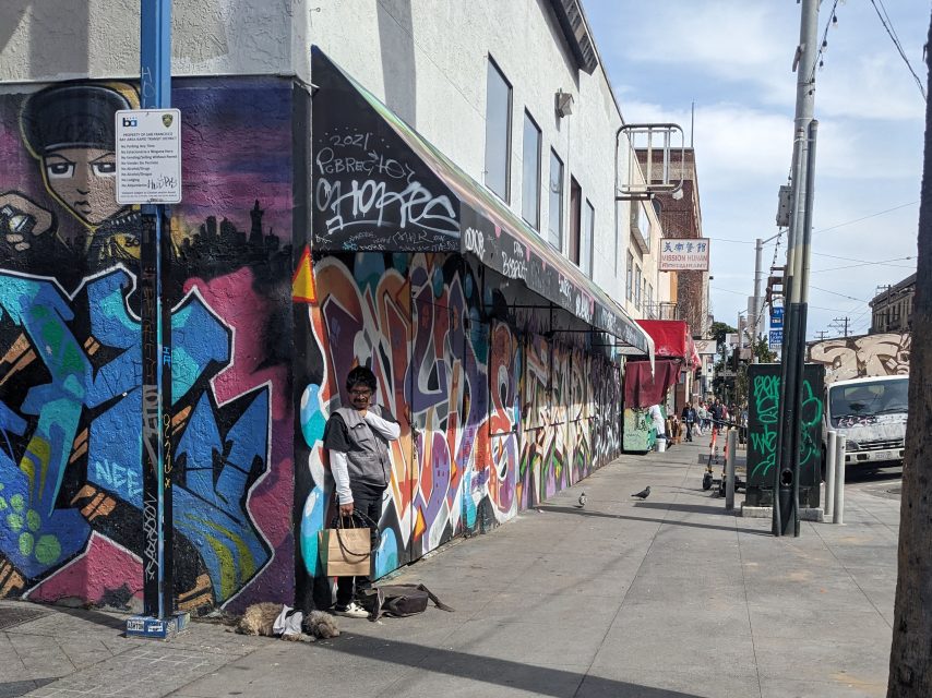 A person stands with his dog against a graffiti-covered corner of a building on an urban sidewalk, with pigeons nearby and people in the distance.