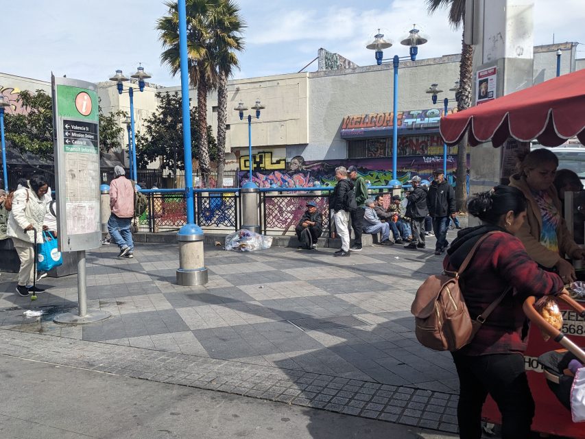 Urban scene with people walking and sitting near street vendors. A fence with graffiti and a palm tree are in the background.