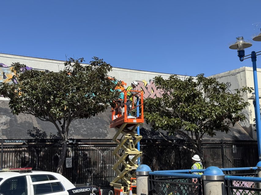 Workers on an orange scissor lift work on graffiti on a wall beside trees. A parked emergency vehicle and street lamp are nearby under a clear blue sky.