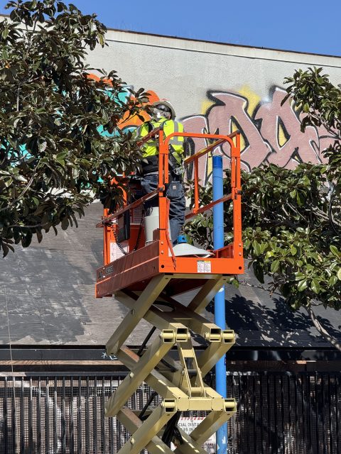 Person in a high-visibility vest standing on a scissor lift, working on graffiti art on a building wall surrounded by leafy branches.