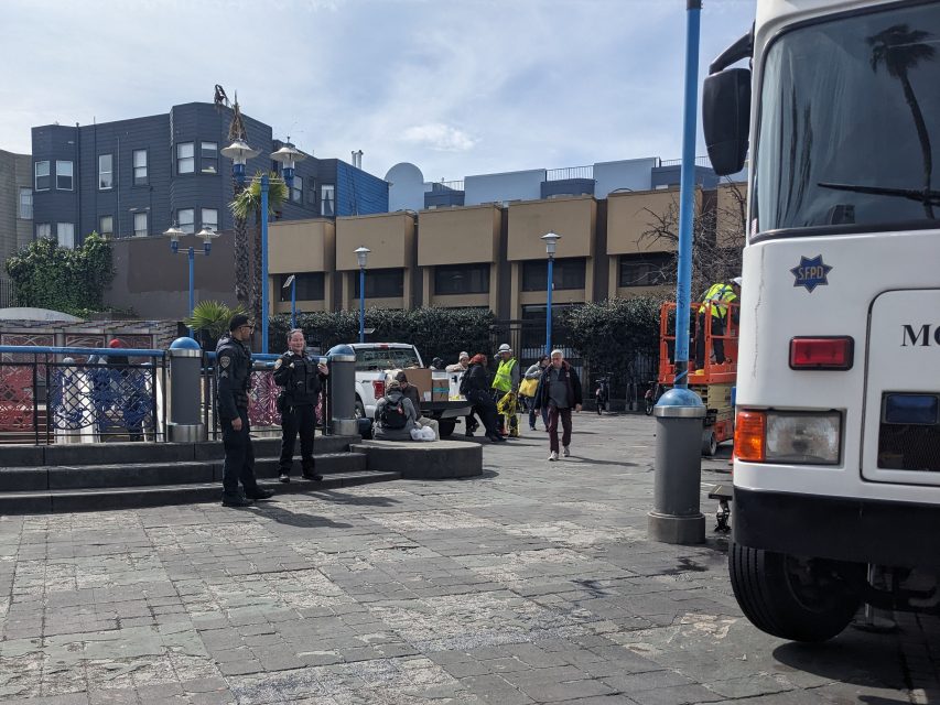 Police officers and workers are present in an urban plaza with a parked utility truck and an orange lift.