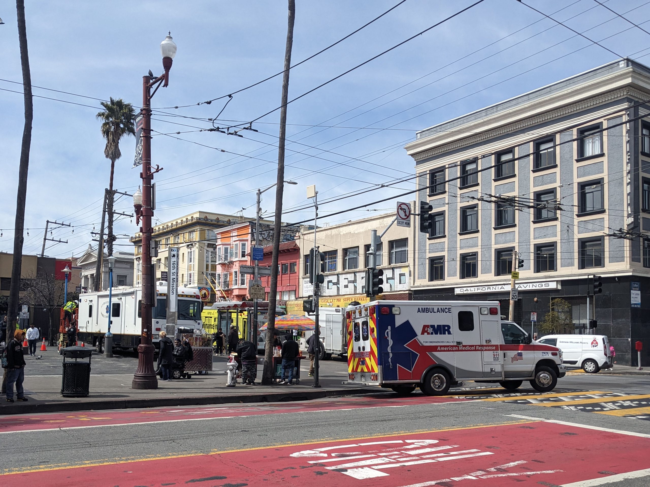 Street scene with an ambulance parked at an intersection, people standing nearby, and buildings in the background. Wires cross the sky, with a palm tree and streetlights visible.
