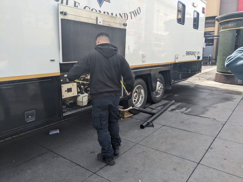 A man in a black hoodie and cargo pants standing beside an open compartment of a large emergency vehicle on the street.