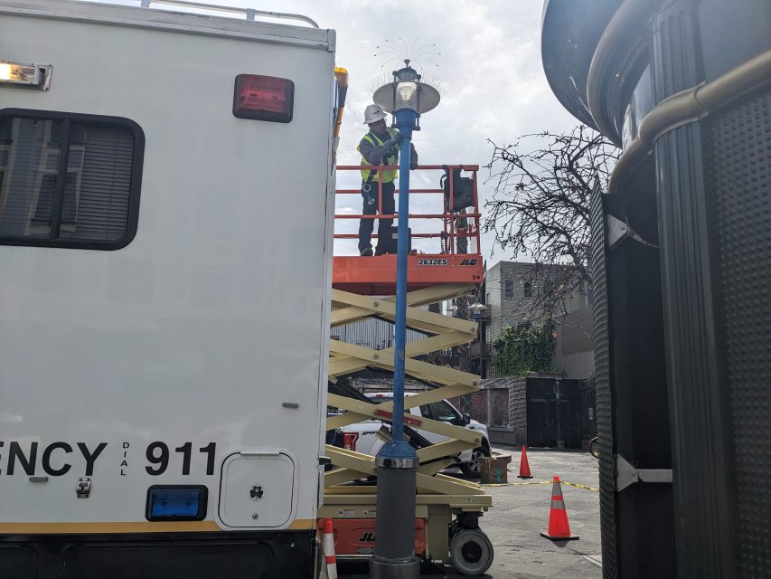 Workers in a lift repair a streetlight next to a utility vehicle on an urban street. Orange cones mark the area for safety.