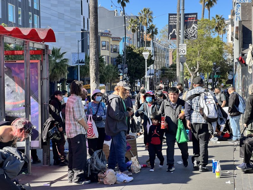 A crowded urban street at the 16th Street BART plaza features people gathered, carrying bags and wearing casual clothing. Some wear masks as urban buildings and palm trees frame the bustling scene.