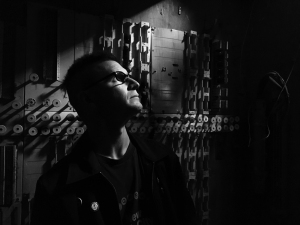 A person wearing glasses looks upward in a dimly lit room with industrial elements on the wall.