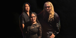Three individuals dressed in black stand against a dark background. The person in front has braided hair, the person on the left has long dark hair, and the person on the right has long blond hair and sunglasses.