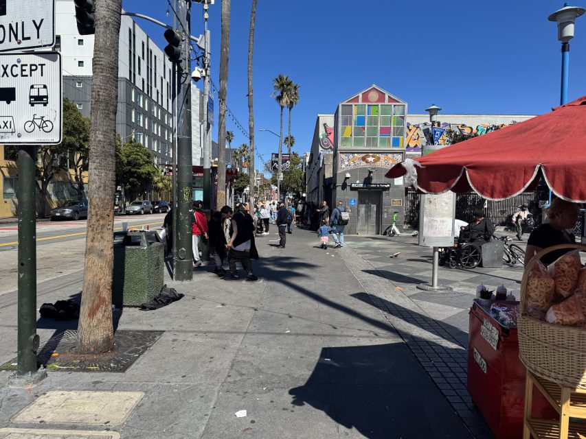 Street scene with pedestrians near a colorful building and food stand. Signage, palm trees, and a red canopy are visible, along with a clear blue sky.