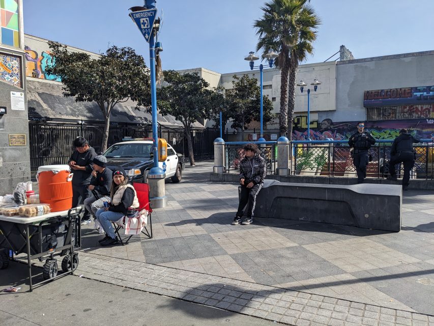 Urban scene with people sitting and standing near a mural and vendor cart in a plaza. A police car is parked nearby, and trees line the area under a blue sky.