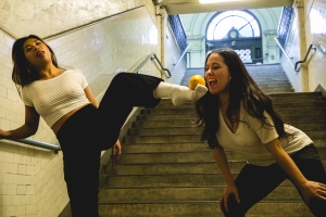 Two women are on a staircase; one is playfully kicking near the other's head. Both appear to be having fun, shown by their expressions and relaxed body language.