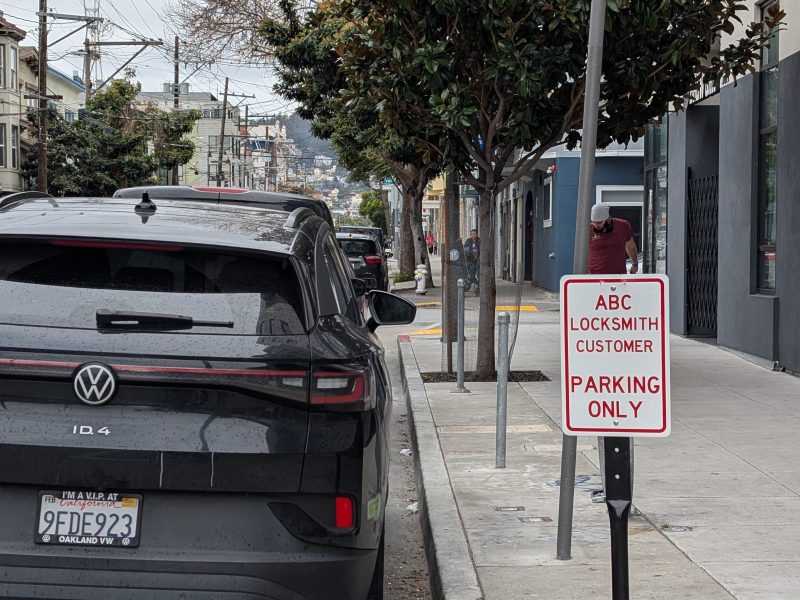 A black Volkswagen ID.4 is parked by the curb near a sign that says "ABC Locksmith Customer Parking Only" on a city street, with trees and a building in the background.
