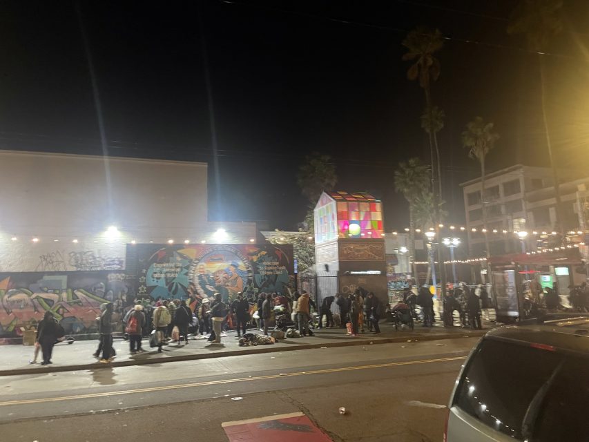 A nighttime street scene shows people in a long line outside buildings with colorful graffiti. String lights are overhead, and palm trees are visible in the background.