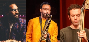 Three musicians: Left is a man with a beard holding a percussion instrument, center is a man in an orange shirt playing a saxophone, right is a man in a green shirt playing a double bass.