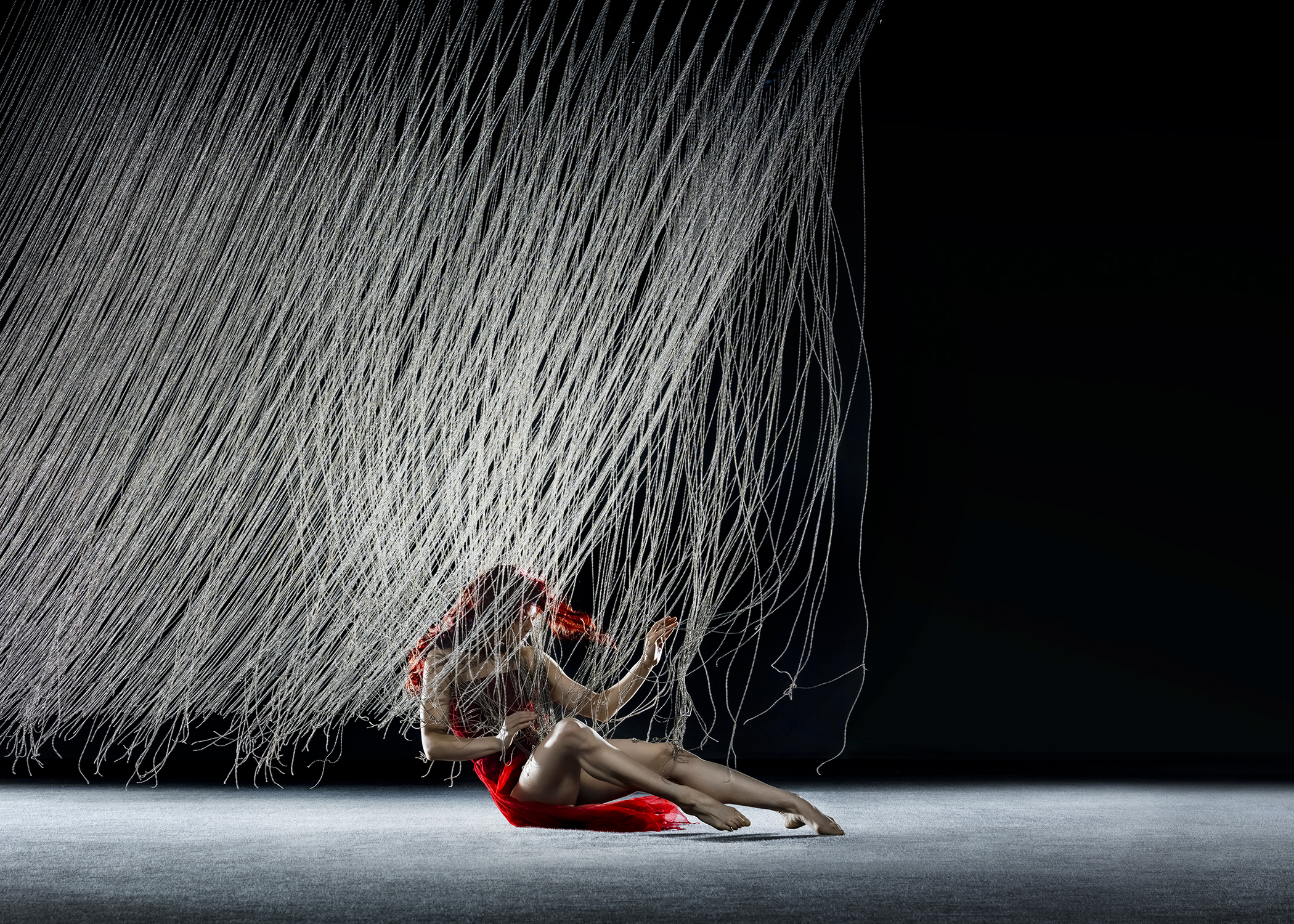 A person in a red dress sits on the floor under a canopy of hanging strings against a dark background.