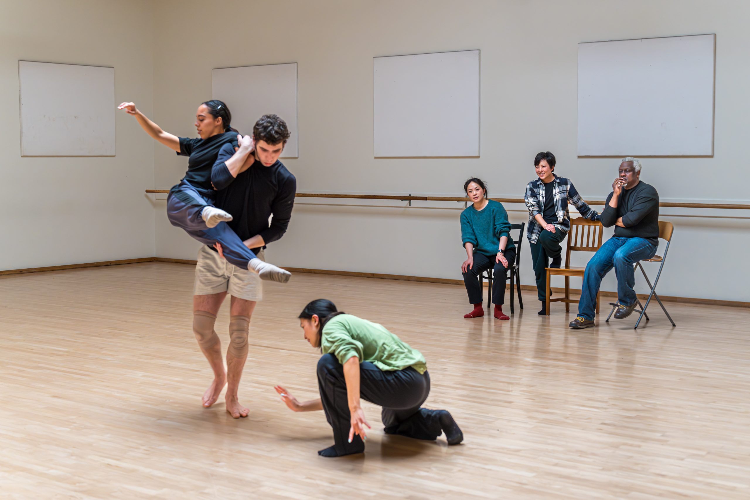 Dancers practicing in a studio, with one performer lifting another. Three people are sitting and watching them from the side.