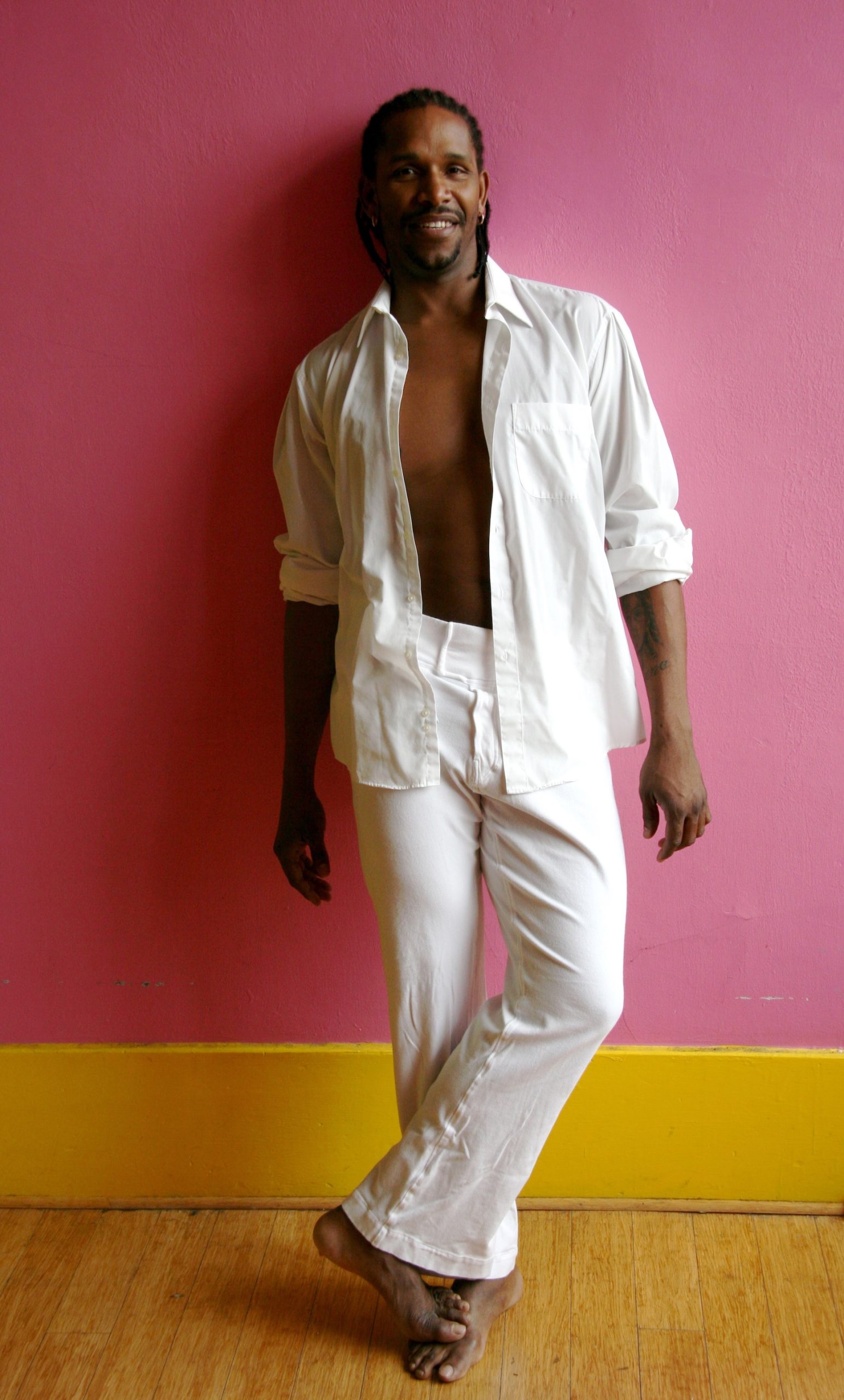 Man in a partially unbuttoned white shirt and pants stands against a pink wall with a yellow base, smiling and barefoot on a wooden floor.