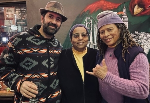 Three people stand closely together, smiling. The person on the left holds a drink. A colorful mural is visible behind them.