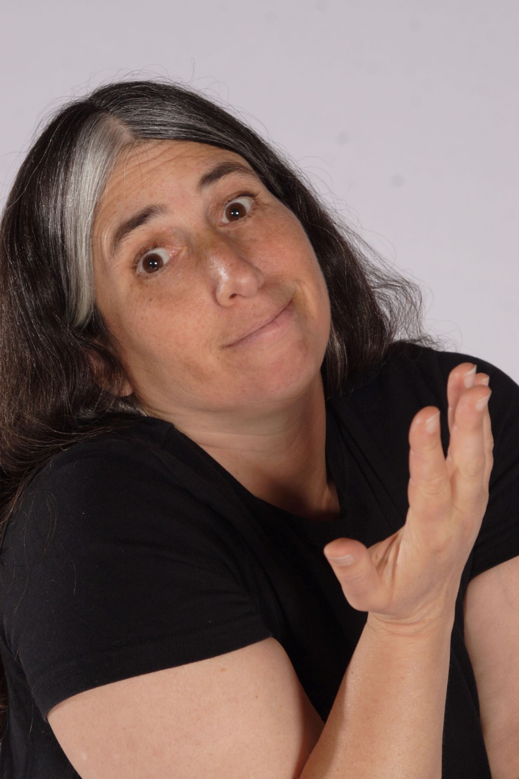 A person with long dark hair and a streak of white shrugs with one hand raised, wearing a black shirt and displaying a neutral expression against a plain background.